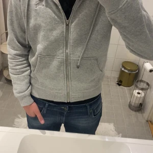 Grå zip hoodie från Burberry - Säljer en grå zip hoodie från Burberry med klassiskt rutigt foder i huvan. Hoodien har dragkedja framtill, två fickor och justerbara snören i huvan. Tillverkad i mjukt bomullsmaterial och har en avslappnad passform. Perfekt för en casual och stilren look.PRIS KAN DISKUTERAS 