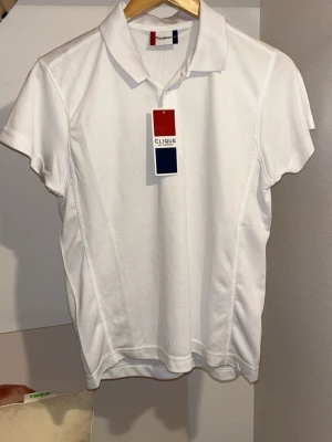 Vit pikétopp från Clique - Klassisk vit pikétopp från Clique med krage och korta ärmar. Tillverkad i ett lätt och bekvämt material som passar perfekt till en sportig stil. Enkel design utan mönster och med diskreta sömmar längs sidorna.