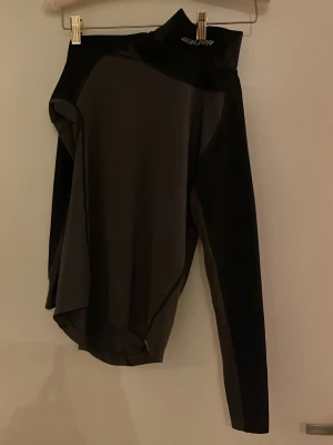 Svart långärmad träningströja Bauer - Svart långärmad träningströja från Bauer med hög krage och diskret logga på axeln. Tröjan är tillverkad i ett stretchigt och funktionsmaterial som andas, perfekt för intensiva sportaktiviteter. Snygg och sportig design med paneler i olika svarta nyanser.