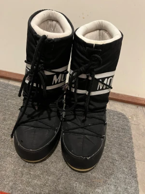 Svarta Moon Boot snowboots - Säljer ett par klassiska svarta Moon Boot snowboots med vit logga och snörning framtill. Bootsens ovandel är i nylon och insidan är fodrad för extra värme. Tjock sula och hög modell som ger retrovibbar och håller dig varm hela vintern. Ena snörning är trasig och ett snöre är utdraget ur resåren. Men inget som stör och går att fixa med nytt snöre.