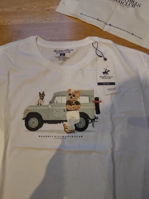 Vit t-shirt Beverly Hills Polo Club - Vit t-shirt från Beverly Hills Polo Club med tryck av en nallebjörn i solglasögon och hund framför en bil. Klassisk passform och rund hals. Perfekt för dig som gillar coola och lekfulla prints.