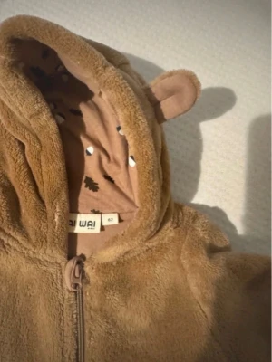Brun teddy jumpsuit med öron - Supersöt brun teddy jumpsuit från Wai Wai med huva som har små björnöron. Dragkedja framtill och mjukt foder med gulligt mönster på insidan av huvan. Perfekt för att hålla sig varm och mysig under kyliga dagar.