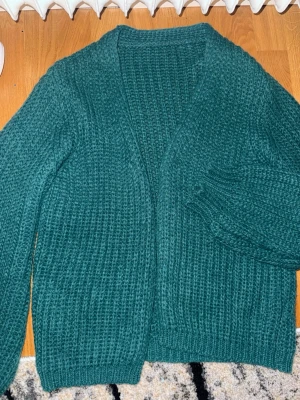 Grön stickad kofta i chunky stil - Mysig grön stickad kofta med grovstickad struktur och avslappnad passform. Koftan har långa ärmar, ribbade muddar och öppen front utan knappar. Perfekt för lager på lager och ger en chill vibe till din outfit. Materialet känns mjukt och värmande.
