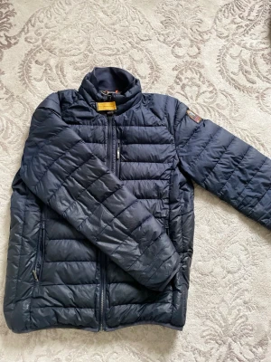 Mörkblå pufferjacka från Parajumpers - Säljer en mörkblå pufferjacka från Parajumpers. Jackan har hög krage, dragkedja framtill och flera praktiska fickor. Quiltad design med klassisk logga på ärmen ger en stilren look – perfekt för våren. I bra begagnat skick med mindre slitningar som knappt märks. Ett mycket litet hål finns baktill. Remmarna i innerfickan har lossnat men fickan fungerar. Priset är satt därefter – ett riktigt bra fynd!