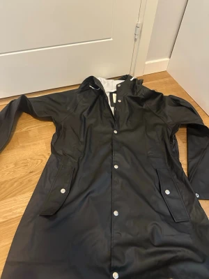 Svart regnjacka från lager  157 - Svart regnjacka från lager 157 med huva och tryckknappar framtill. Jackan har två stora fickor med lock och vita detaljer på insidan. Materialet är vattenavvisande och perfekt för regniga dagar. Enkel och stilren design som passar till det mesta.