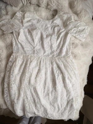 Vit spetsklänning med kort ärm - Superfin vit kortklänning i spets med rund halsringning och korta ärmar. Klänningen har en lätt utställd kjol och vackra spetsdetaljer över hela plagget. Perfekt för dig som gillar romantisk och feminin stil.