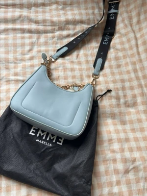 Ljusblå handväska från Emme Marella - Trendig ljusblå handväska från Emme Marella i slätt konstläder. Väskan har guldfärgade detaljer, avtagbar axelrem med logotyp och en kedja. Medföljer matchande kortfodral och dustbag. Perfekt accessoar för att lyfta din outfit.