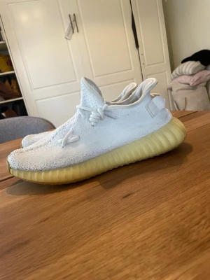 Vita Yeezy Boost 350 V2 sneakers - Säljer ett par vita Yeezy Boost 350 V2 från Adidas. Skorna har en stickad ovandel i Primeknit, vita snören och en tjock, ribbad sula i gulbeige ton. Ikonisk siluett med bekväm passform och modern design. Perfekta för dig som gillar streetwear och sneakers med hype.