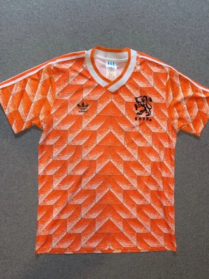 Holländsk retro fotbollströja Adidas - Säljer en ikonisk orange fotbollströja från Adidas med KNVB-logga och svart lejon på bröstet. Tröjan har ett retro geometriskt mönster, vita ränder på axlarna och V-ringad krage. Perfekt för dig som älskar klassiska fotbollströjor och vill sticka ut på planen. Använt en gång 