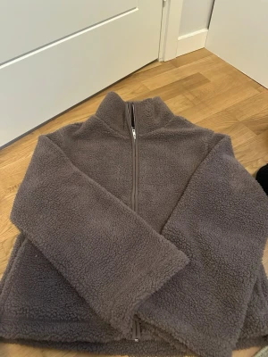 Brun teddyjacka med dragkedja - Mysig brun teddyjacka med hög krage och hel dragkedja framtill. Jackan har en avslappnad passform och är tillverkad i fluffigt teddy-material som håller dig varm. Perfekt för lager-på-lager och chill dagar.