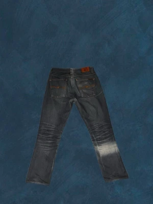 Mörkblå jeans från Nudie Jeans - Snygga mörkblå jeans från Nudie Jeans med raka/baggie ben och klassiska bakfickor. Jeansen har slitningar och ljusare partier på benen för en cool, använd look. Tillverkade i denim med kontrastsömmar och läderpatch bak i midjan. Jeansen är professionell uppsydda från W32/L32-W32/L30~