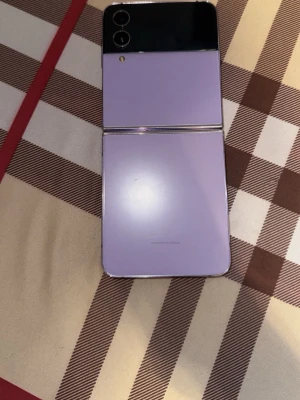 Samsung Galaxy Z Flip lila - Snygg vikbar Samsung Galaxy Z Flip i lila färg med glasbaksida och dubbla kameror på ovansidan. Telefonen har en modern vikbar design och stor skärm på insidan. Perfekt för dig som vill ha en unik och smidig Android-mobil.