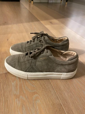 Gröna mockasneakers från ARIGATO - Stilrena sneakers i grön mocka från ARIGATO med vita sulor och klassisk låg profil. Skorna har snörning och diskret logga i guld på sidan. Perfekta för dig som gillar minimalistisk och clean stil.