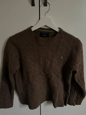 Brun kabelstickad tröja från GANT - Mysig brun kabelstickad tröja från GANT i 100% lammull. Tröjan har rund halsringning, ribbade muddar och ett broderat GANT-emblem på bröstet. Perfekt för dig som gillar klassisk och tidlös stil.
