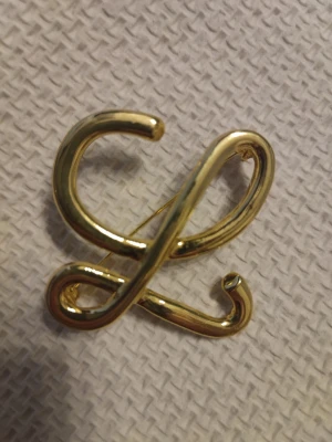 Guldfärgad brosch i form av bokstav - Snygg brosch i guldfärgad metall, formad som en stiliserad bokstav eller symbol. Perfekt att fästa på jackan eller väskan för en personlig touch. Enkel nål på baksidan för smidig användning.