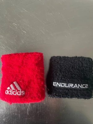Adidas & Endurance svettband - Två sportiga svettband, ett rött från Adidas med broderad logga och ett svart från Endurance med vit text. Perfekta för träning och sportaktiviteter. Tillverkade i mjukt och absorberande frottématerial.