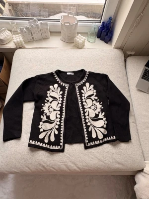 Svart kofta med vit brodyr - Säljer en svart kofta med långa ärmar och öppen front. Koftan har detaljerad vit brodyr i blommigt mönster framtill och dekorativa kanter. Perfekt för dig som gillar unika plagg med retrovibbar och snygga kontraster. Koftan är köpt från en spansk marknad i mallorca!