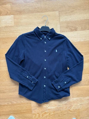 Mörkblå skjorta från Ralph Lauren - Klassisk mörkblå skjorta från Ralph Lauren med button-down krage och broderad logga på bröstet. Skjortan har långa ärmar, vita knappar och är tillverkad i mjuk bomull. Perfekt för en stilren och avslappnad look. Knapppats andvänd, säljer på grund av att jag växt ur den.Tveka inte på att höra av vid eventuella frågor!
