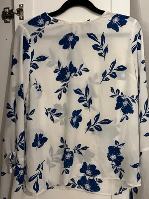 Vit blommig blus med blått mönster - Säljer en luftig vit blus med blåa blommor och blad. Blusen har långa ärmar med knytband vid ärmsluten och en rak, avslappnad passform. Materialet känns lätt och flowy, perfekt för en stilren look. ⭐️Tröjan är i bra skick förutom att det verkar vara någon tråd som ligger löst vid knappen bak (se bild)🩷 