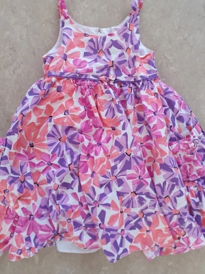 Klänning - Söt blommig klänning från Maggie & Zoe i färgerna rosa, lila och orange. Klänningen har smala axelband och ett luftigt, utställt snitt. Tillverkad i bomull med foder i polyester och bomull. Perfekt för soliga dagar!