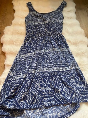 Blåvit mönstrad sommarklänning - Snygg blåvit klänning med grafiskt mönster och breda axelband. Klänningen har en avslappnad passform och är tillverkad i ett mjukt, lätt material som passar perfekt för varma dagar. Midjan är lätt markerad och kjolen har en luftig känsla.