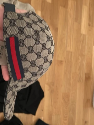 Gucci keps med GG-mönster - Säljer en snygg beige keps från Gucci med klassiskt GG-monogram över hela kepsen och en ikonisk röd och marinblå rand på sidan. Kepsen har böjd skärm och justerbar rem bak. Perfekt för dig som vill ha en stilren och lyxig accessoar.