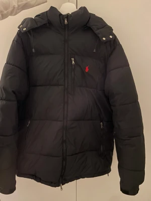 Svart pufferjacka från Polo Ralph Lauren - Snygg svart pufferjacka från Polo Ralph Lauren med röd logga på bröstet. Jackan har huva med tryckknappar, dragkedja framtill och flera fickor med dragkedja. Perfekt för kalla vinterdagar och riktigt skön att bära.