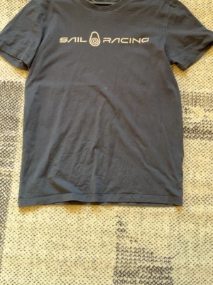 Mörkblå Sail Racing t-shirt - Snygg mörkblå t-shirt från Sail Racing med vit logga tryckt över bröstet. Klassisk passform och rund halsringning. Tillverkad i mjuk bomull som känns skön mot huden. Perfekt för en avslappnad och stilren look.