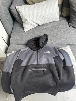 The North Face svart/grå hoodie - Säljer en svart och grå hoodie från The North Face med halv dragkedja och stor ficka framtill med dragkedja. Hoodien har en klassisk huva och är tillverkad i mjukt material, perfekt för chill dagar eller när du vill ha en sportig look.