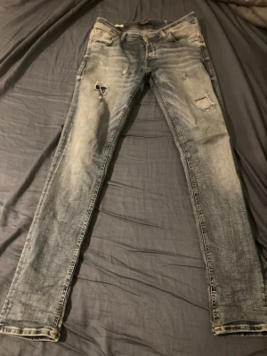 J&J Slim fit  blå jeans med slitningar  - Säljer ett par slim fit blå jeans med slitningar och hål på låret och knät. Jeansen har klassisk femficksdesign, dragkedjegylf och är tillverkade i denim. Från Jack and Jones modellen är slim fit glen. Storlek W31 L34, Pris kan diskuteras vid snabb affär 