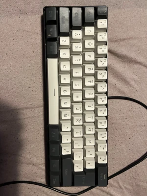 Kompakt mekaniskt tangentbord RGB - Säljer ett kompakt mekaniskt tangentbord med svart och vita tangenter. Tangentbordet har RGB-belysning som lyser i flera färger och ansluts med kabel. Perfekt för gaming eller skrivbordssetup där du vill spara plats.