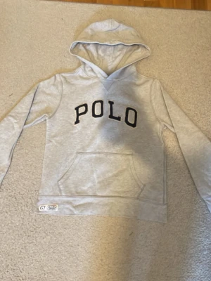 Grå hoodie från Polo Ralph Lauren - Snygg grå hoodie från Polo Ralph Lauren med svart POLO-tryck på bröstet och en klassisk känguruficka. Tröjan har huva och en liten etikett med '67' och Polo RL-detaljer vid nederkanten. Perfekt för en chill och sportig look.