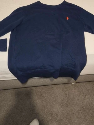 Mörkblå stickad tröja från Ralph Lauren - Mörkblå stickad tröja från Ralph Lauren med klassisk rund halsringning och den ikoniska röda logotypen broderad på bröstet. Tröjan har långa ärmar och ribbade muddar vid ärmslut och nederkant. Perfekt för en stilren och avslappnad look.