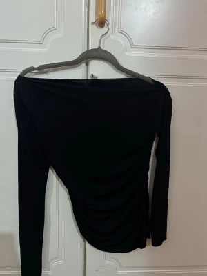 Svart offshoulder topp med rynk - Trendig svart offshoulder topp med långa ärmar och snygga rynkdetaljer på sidan. Tunn och stretchig, perfekt för att styla med höga jeans eller kjol. Enkel men ändå elegant design 