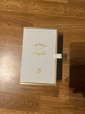 Lattafa The Kingdom Eau de Parfum - Lyxig parfym från Lattafa, The Kingdom, i en elegant guldfärgad glasflaska med räfflad yta. Kommer i en exklusiv vit och guld presentförpackning med gulddetaljer och dekorativ text. Perfekt för dig som gillar exklusiva dofter och snygg design.