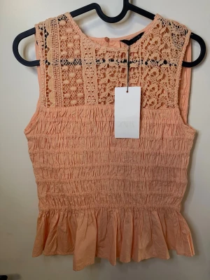 Persikofärgad spetsblus från Zara med prislapp - Supersöt persikofärgad blus från Zara med spetsdetaljer upptill och smockad midja. Ärmlös modell med volangkant nedtill och vackert broderat mönster på axlar och rygg. Perfekt för dig som gillar romantisk och feminin stil.