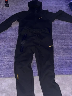 Storlek s passar m. ocksåSvart Nike Tech Fleece set - Storlek s passar m också. Nike Tech Fleece set i svart med guldfärgade detaljer. Setet består av en zip-hoodie med hög krage och två fickor samt matchande byxor med dragsko och fickor. Både jackan och byxorna har Nike-logga i gult och byxorna har 'NOCTA' tryckt på benet. Perfekt för chill eller streetwear.