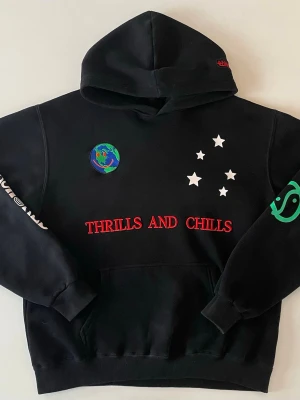 Svart hoodie Thrills and Chills - PRISET GÅR ATT DISKUTERA.                Svart hoodie med huva och coola tryck som 'THRILLS AND CHILLS' i rött framtill, vita stjärnor, jordglob och grönt motiv på ärmen. Baksidan har blå text 'OUT OF THIS WORLD', vit text och ett stort rött peace-märke. Perfekt för dig som gillar streetwear och unika detaljer.