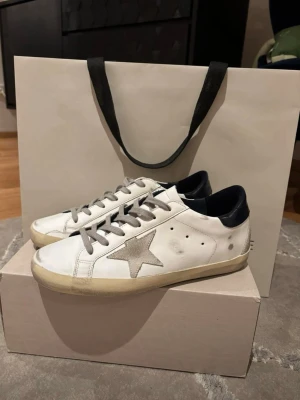 Golden Goose vita sneakers med stjärna - Golden Goose sneakers i vitt läder med grå stjärndetalj på sidan, grå skosnören och svart hälparti. Klassisk låg modell med slitna detaljer för en cool vintage-look. Yttersula i gummi med fiskbensmönster. Perfekta för dig som gillar streetstyle.