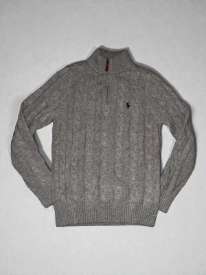 Grå kabelstickad tröja Polo Ralph Lauren - Kabelstickad grå tröja från Polo Ralph Lauren med halv dragkedja och hög krage. Tröjan har ribbade muddar och nederkant samt den klassiska broderade loggan på bröstet. Perfekt för lager på lager och en stilren look.