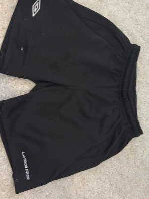 Svarta shorts från Umbro - Svarta sportiga shorts från Umbro med elastisk midja och vitt logotryck på benet. Lätta och bekväma med meshfoder på insidan, perfekta för fotboll eller träning.
