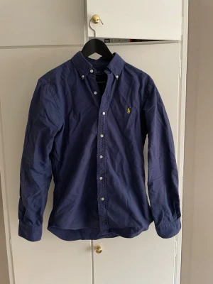 Skjorta från Ralph Lauren - Klassisk marinblå skjorta från Ralph Lauren med button-down krage och broderad gul logga på bröstet. Den är tillverkad i bomull och är i storlek S. Den är lite skrynklig på bilderna men löser sig när man stryker den. Tveka inte på att höra av dig om du har några frågor.