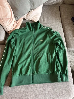 Grön ziptröja från Lacoste - Snygg grön ziptröja från Lacoste med regular fit. Tröjan har dragkedja framtill, ribbade muddar och vita detaljer längs ärmarna. Klassisk Lacoste-logga på bröstet. Perfekt till streetwear eller sportig stil.