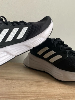 Svarta Adidas med vita ränder - Snygga svarta Adidas sneakers med klassiska vita ränder på sidorna och vit, chunky sula. Ovandelen är i meshmaterial som andas och skorna har svarta skosnören. Perfekta för en sportig och stilren look. Använd ett par gånger inomhus, som nya.