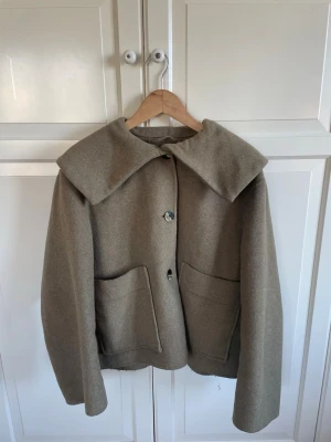 Beige ulljacka från Ganni - Stilren beige jacka från Ganni i ull med stor krage och två rejäla framfickor. Jackan har knäppning framtill och en rak, något boxig passform. Perfekt för dig som gillar minimalistisk och trendig design. Den har några ”fläckar” vid ena axeln men ingenting som syns tydligt 