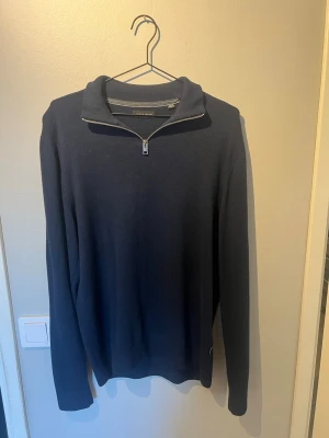 Mörkblå halvzip-tröja - Mörkblå stickad tröja med halvzip och hög krage. Tröjan har en stilren design med långa ärmar och ribbade muddar. Perfekt för lager på lager och enkel att matcha med jeans eller chinos.