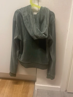 Grön velour hoodie med dragkedja - Mjuk grön hoodie i velour med dragkedja framtill och känguruficka. Tröjan har huva med snören och ribbade muddar vid ärmslut och nederkant. Liten broderad logga på bröstet ger en snygg detalj. Perfekt för en chill och avslappnad stil.