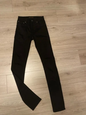 Svarta skinny jeans från Nudie Jeans - Svarta skinny jeans från Nudie Jeans med klassisk femficksdesign och silverfärgade nitar. Jeansen har en smal passform och är tillverkade i stretchigt denimtyg för extra komfort. Perfekta för en stilren och modern look.