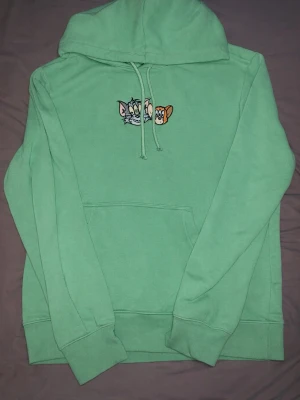Grön hoodie med Tom & Jerry brodyr - Snygg mintgrön hoodie med broderade Tom & Jerry-figurer på bröstet. Klassisk känguruficka framtill, ribbade muddar och justerbar huva med dragsko. Perfekt för dig som gillar retro vibes och tecknade serier. Mjuk bomullsblandning för skön känsla.