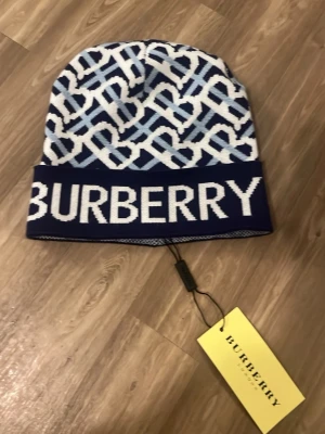 Blåvit Burberry mössa med logga - Snygg blåvit mössa från Burberry med stort logotyp-tryck framtill och grafiskt mönster i vitt och ljusblått. Tillverkad i mjukt stickat material som känns lyxigt och trendigt. Perfekt accessoar för dig som vill sticka ut med en exklusiv look.